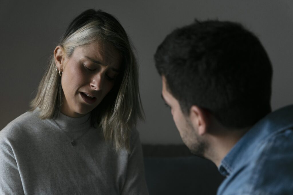A woman appears emotionally distressed while speaking to a man, who listens with concern, in a dimly lit room.